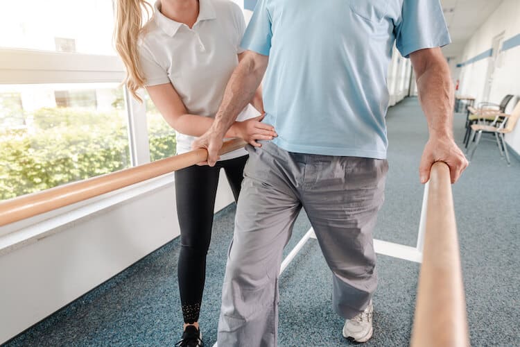 A senior man being helped along walking bars by a physical therapist at a senior rehab center.